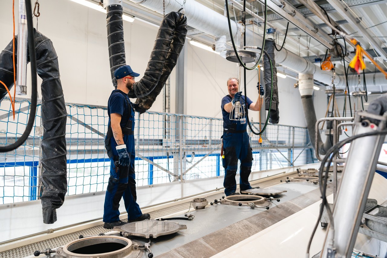 Zwei Arbeiter in blauen Overalls und Mützen auf einem Tankwagen, die Schläuche in offene Luken einführen.