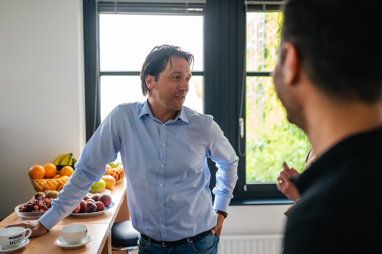 Ein Mann in einem blauen Hemd lehnt an einem Tisch mit Obst und Tassen und spricht mit einer anderen Person.