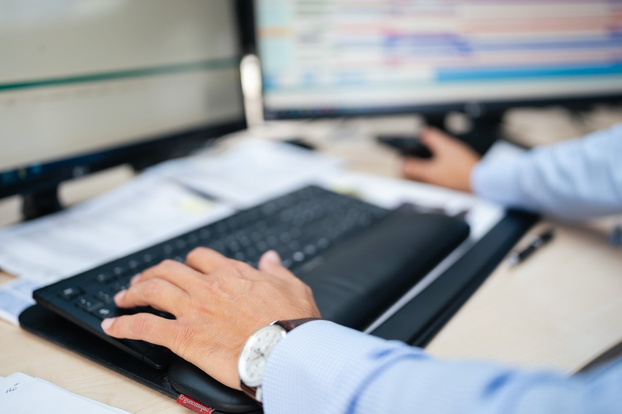 Eine Person tippt an einer Tastatur mit Handgelenkauflage, während ein Computerbildschirm im Hintergrund unscharf zu sehen ist.