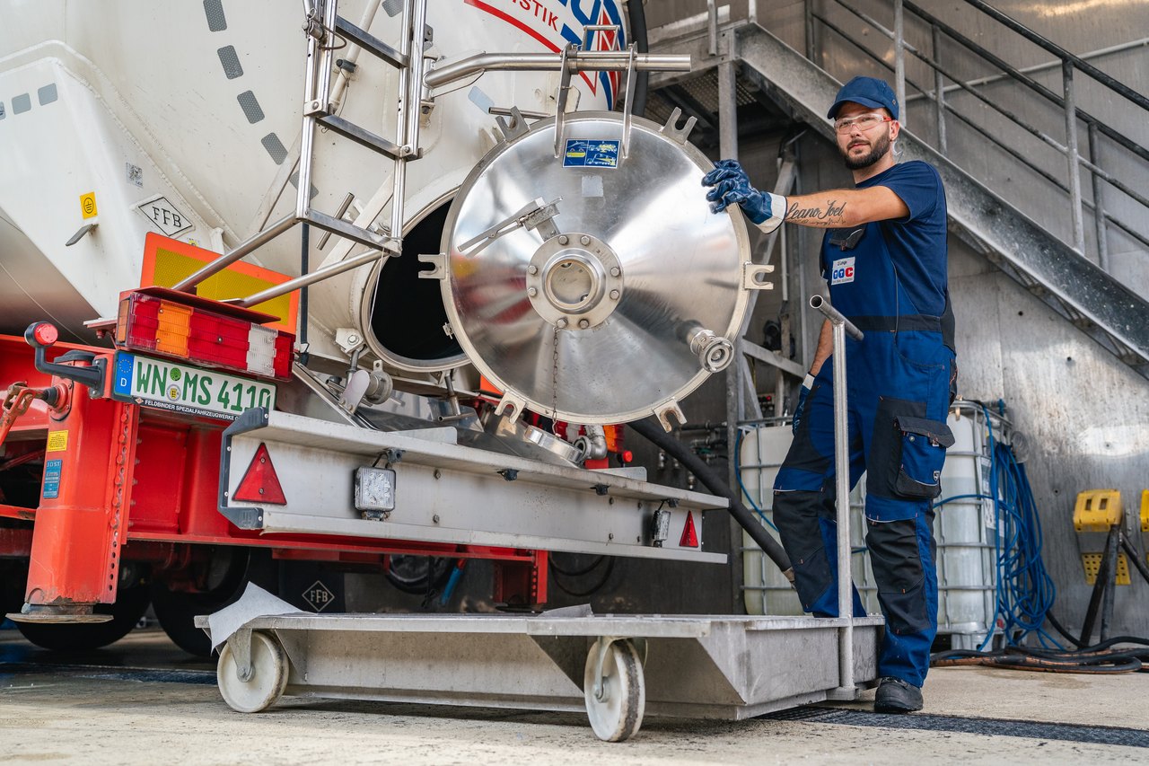 Ein Mann in Arbeitskleidung öffnet einen großen, runden Tankdeckel an einem weißen Tanklastwagen.