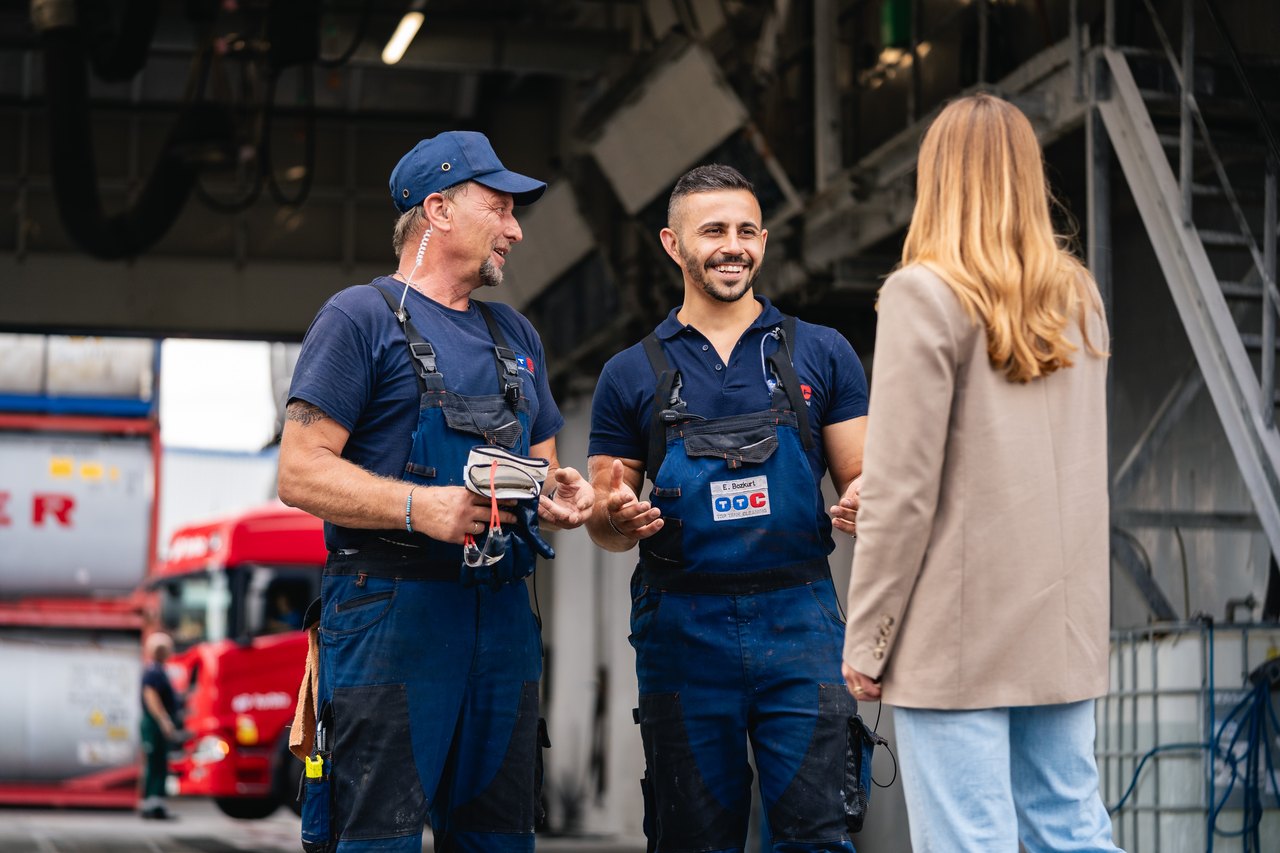 Zwei Arbeiter in blauen Overalls sprechen mit einer Frau im Gebäude, Lastwagen im Hintergrund.
