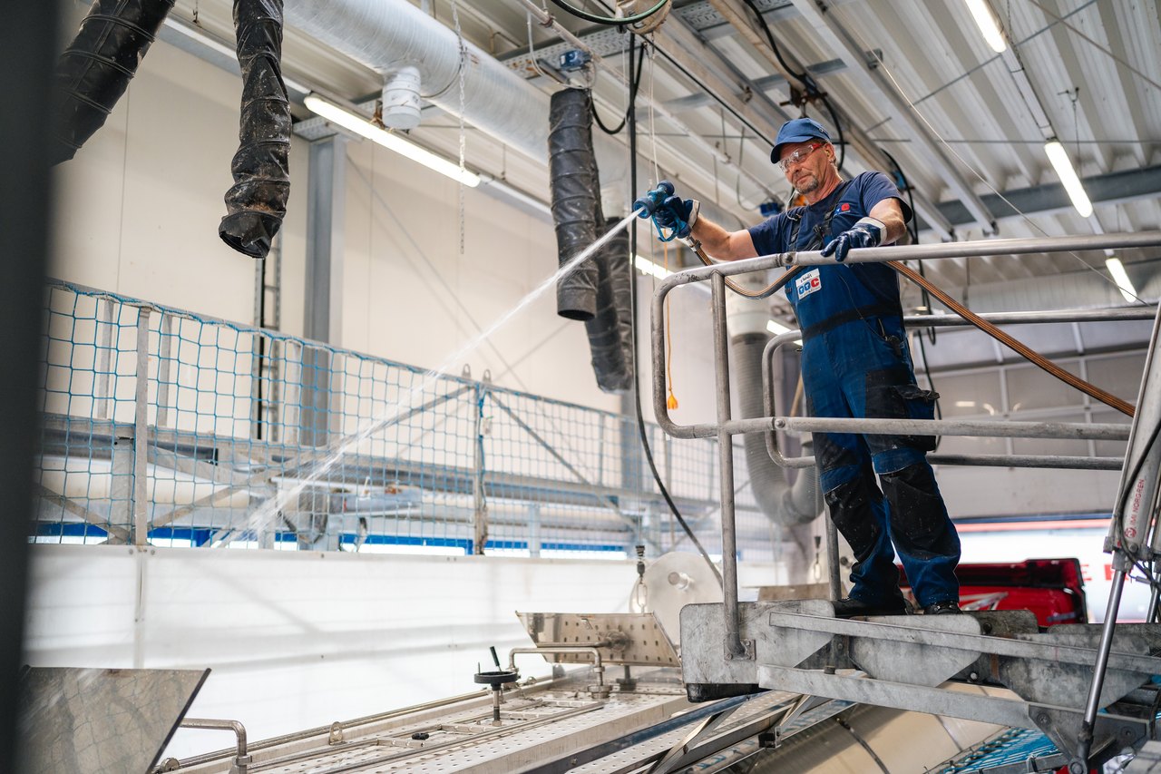 Arbeiter in blauer Uniform und Mütze sprüht Wasser aus einem Schlauch auf große Metalltanks in einer Fabrik.