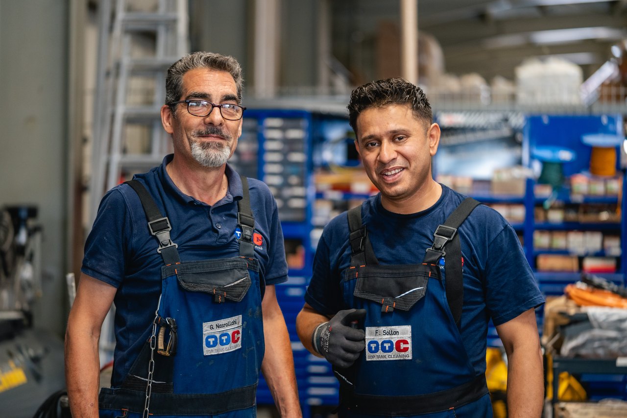 Zwei lächelnde Mechaniker in blauen Overalls mit "OCC"-Logo stehen in einer Werkstatt.
