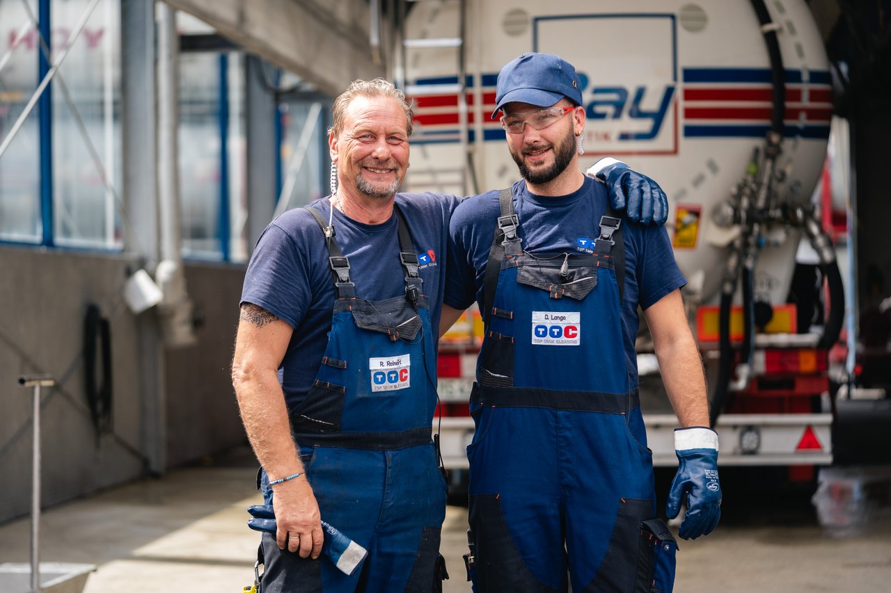 Zwei Männer in blauen Overalls und T-Shirts lächeln, ein Tankwagen mit "bay" Logo im Hintergrund.