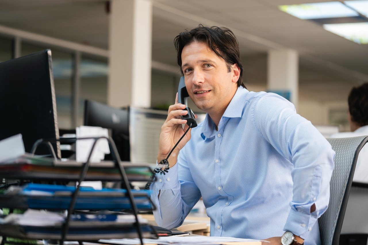 Ein lächelnder Mann in blauem Hemd telefoniert am Schreibtisch im Büro.