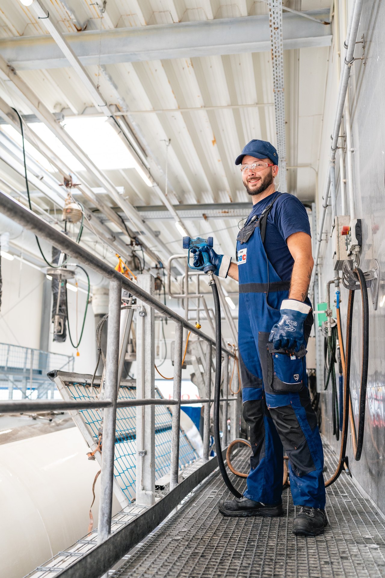 Ein lächelnder Mann in blauer Arbeitskleidung, Schutzbrille und Mütze steht auf einem Gittersteg in einer Fabrik.